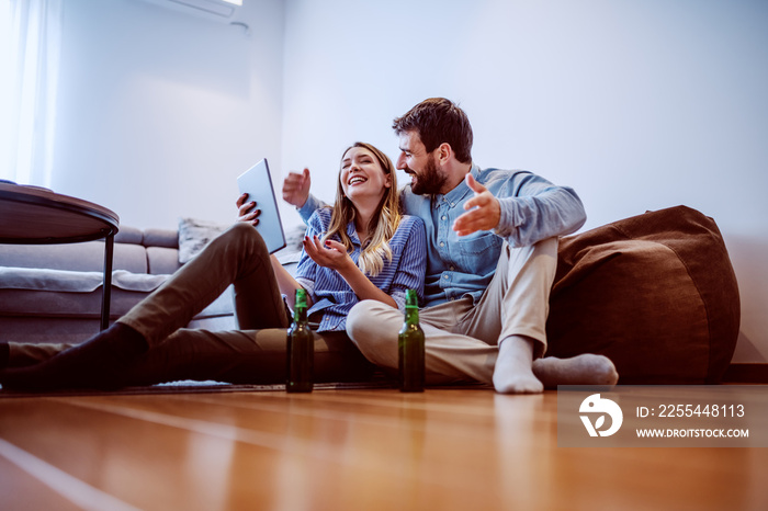 Young cheerful caucasian couple in love sitting on the floor in living room and looking at tablet. On the floor are beer bottles. Free time at sunday afternoon.