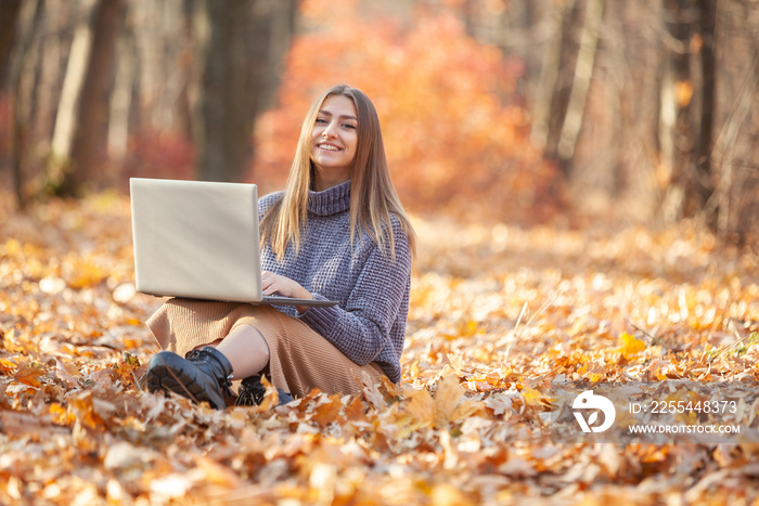 Young female freelancer or student with a laptop sits on fallen leaves in the forest. Remote work or education