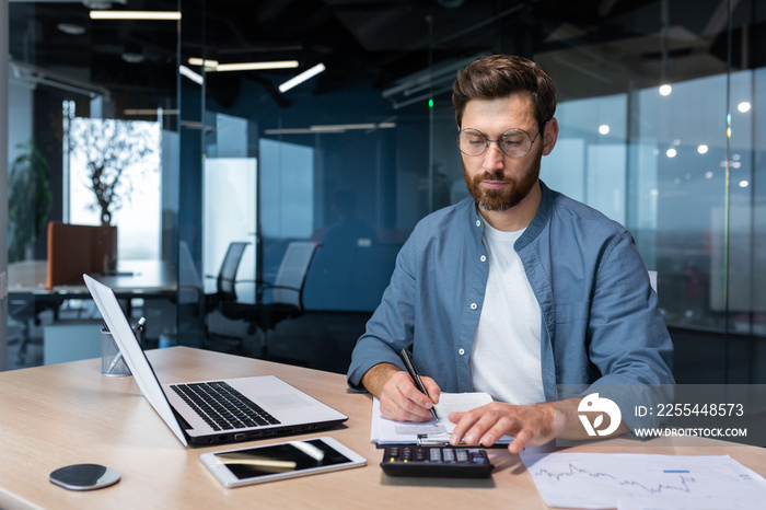 Serious and focused financier accountant on paper work inside office, mature man using calculator and laptop for calculating reports and summarizing accounts, businessman at work in casual clothes.