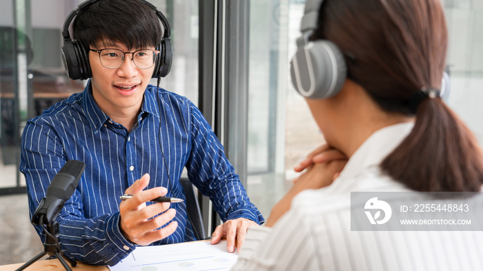 Asian woman radio hosts gesturing to microphone while interviewing a man guest in a studio while recording podcast for online show in studio together