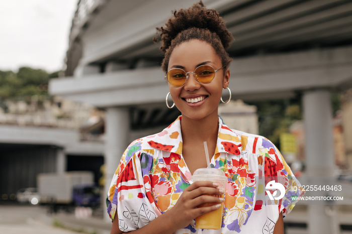 Portrait of curly brunette dark-skinned woman in colorful bright shirt and sunglasses smiles sincerely, holds orange juice glass and poses outside.