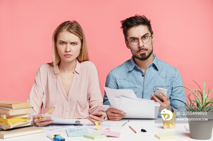 Displeased female and male students sit at work place, study papers together, use modern technologies, have discontent grumpy expressions as have much work to do, isolated over pink background