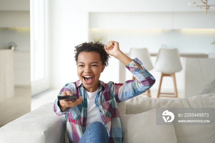 Excited mixed race young girl sport fan watching football on tv, screaming, makes yes winner gesture