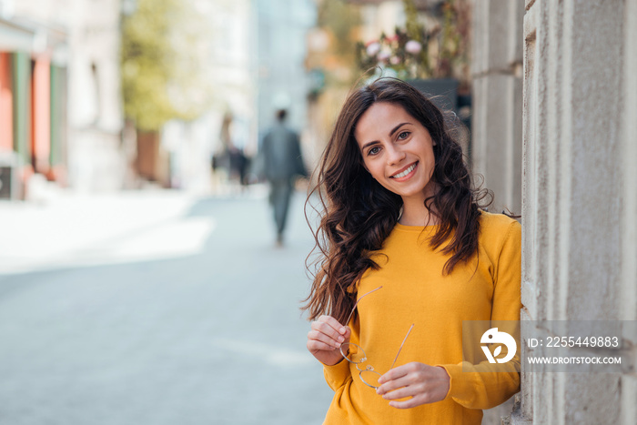 Portrait of a casual young woman in the city street, holding eyeglasses and smiling at camera.