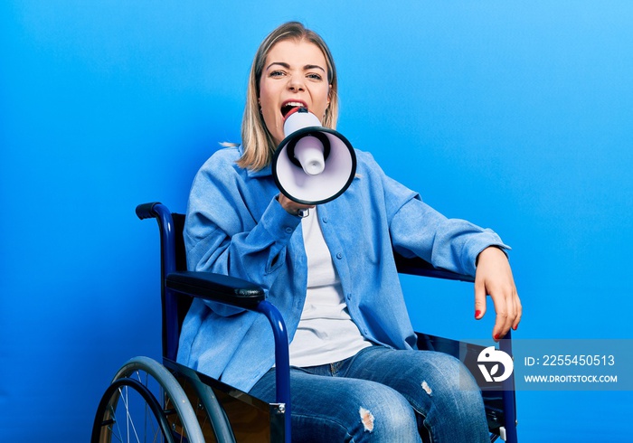 Blonde woman sitting on wheelchair screaming through megaphone over isolated background