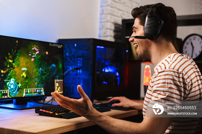 Image of cheerful gamer man playing video games on computer, wearing headphones and using backlit colorful keyboard