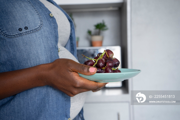 Mid section of pregnant woman holding plate with red grapes in kitchen