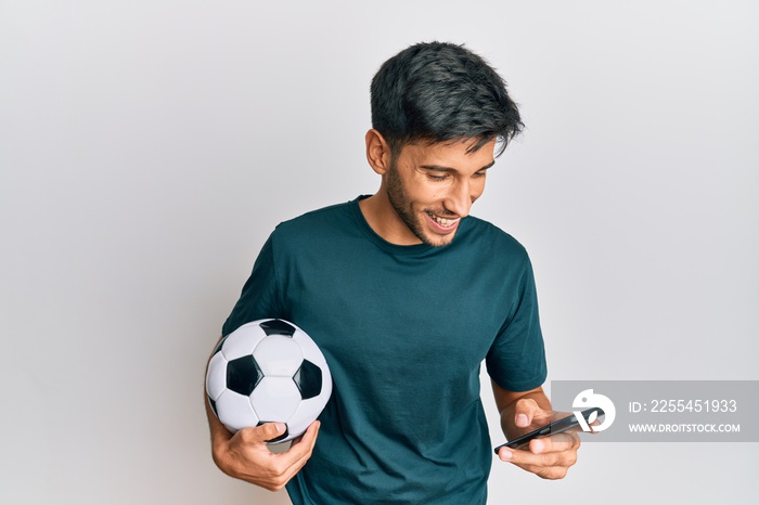 Young handsome man holding football ball looking at smartphone bet app smiling and laughing hard out loud because funny crazy joke.