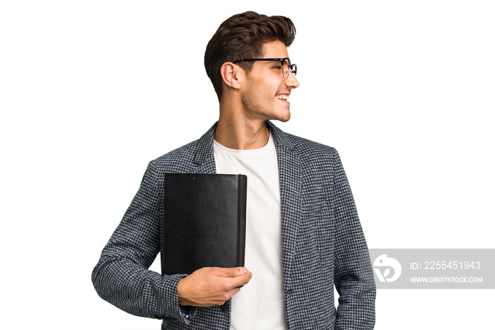 Young teacher caucasian man holding a book isolated looks aside smiling, cheerful and pleasant.