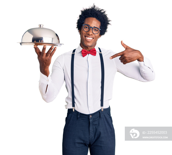 Handsome african american man with afro hair wearing waiter uniform holding silver tray pointing finger to one self smiling happy and proud