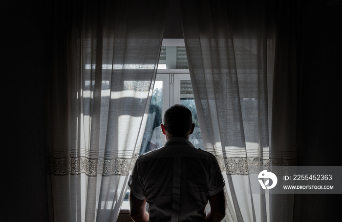 Rear view of adult man standing beside window with sunlight and shadow