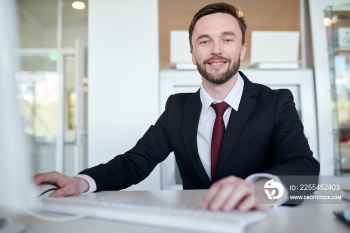 Portrait of bearded handsome businessman posing sitting at desk and looking at camera with charming smile while using computer