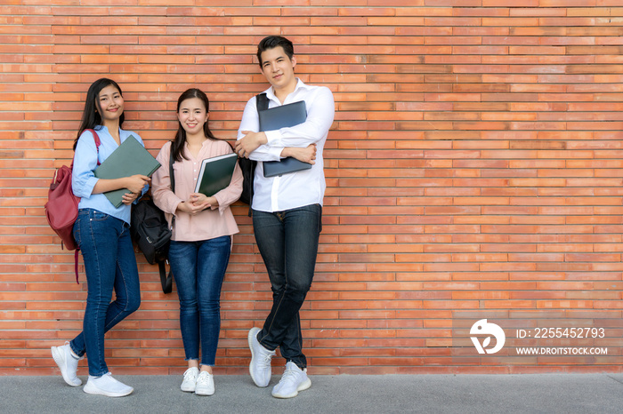 Three Asian smiling student holding book and laptop posing on brick background in campus. Group of happy teen high school students outdoors..