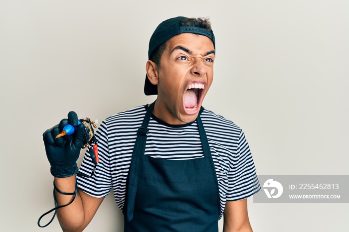 Young handsome african american man tattoo artist wearing professional uniform and gloves holding tattooer machine angry and mad screaming frustrated and furious, shouting with anger