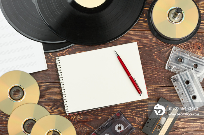 Retro audio devices and blank notebook. Work space with vinyl records, compact discs, analogue cassettes, musical notes, paper notepad and pen. Vintage audio storage.