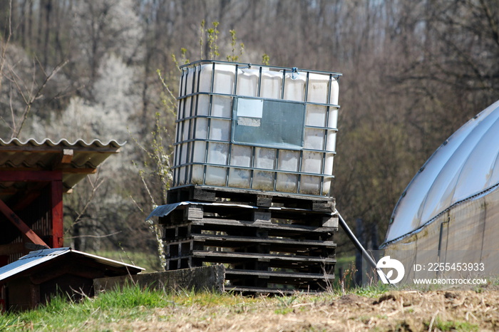 Tilted white intermediate bulk container or IBC plastic tank with metal cage put on top of wooden pallets in local garden to be used for water storage on warm sunny spring day