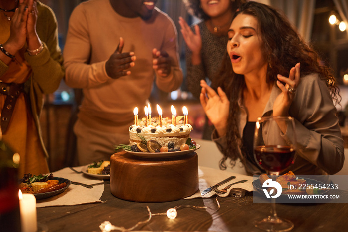 Young woman blowing out candles on birthday cake at the table with her friends clapping hands and congratulating her