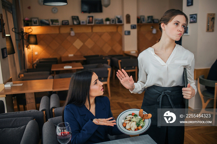 Humble young businesswoman sit at table and show salad bowl to waitress. She refuse to look at it. Young woman in white blouse show stop sign.