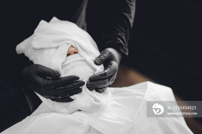 Man sits in barbershop chair and steam his face with hot towel in front of royal beard shaving razor