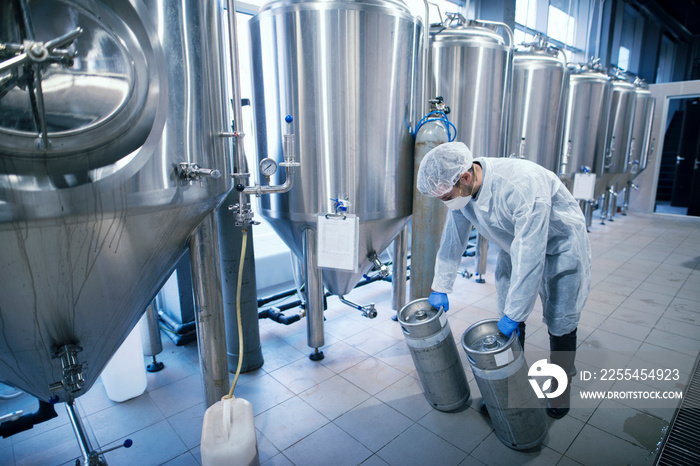 Technologist working in pharmaceutical factory for medical production. Worker in protective suit carrying chemicals in metal reservoirs.