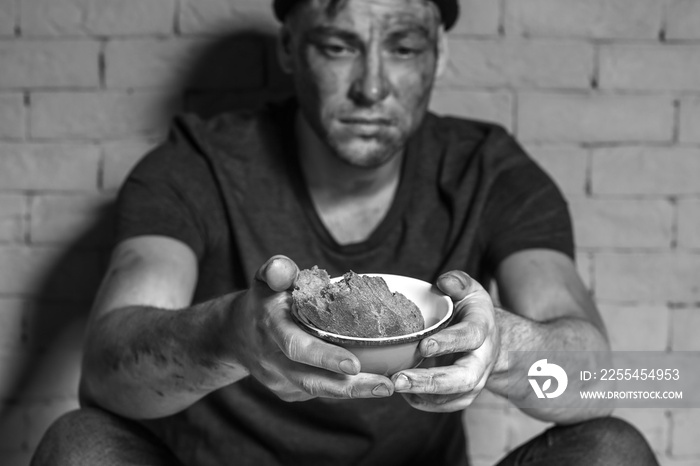 Hungry poor man holding bowl with piece of bread while sitting near brick wall