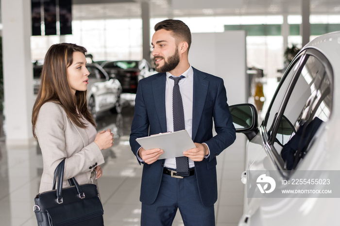 Portrait of handsome car salesman talking to young woman helping her choose luxury car  in showroom