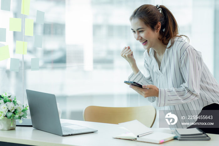 Excited Asian young woman using phone and laptop sitting on a desk office in the day at office