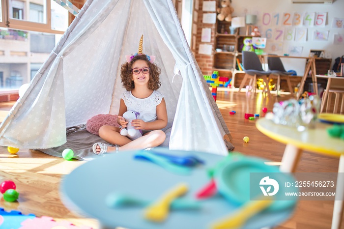 Beautiful toddler wearing glasses and unicorn diadem sitting on the floor inside tipi holding doll and smiling at kindergarten