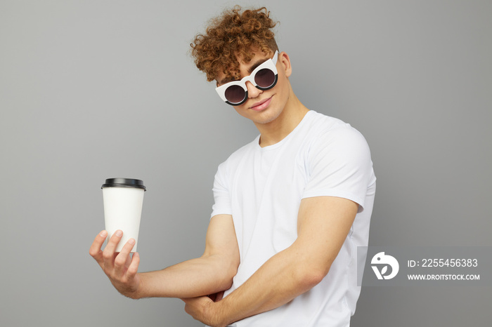 a young man of athletic build stands dressed in a white T-shirt, with sunglasses on his face and holding a white glass in his hand looking at the camera, standing sideways