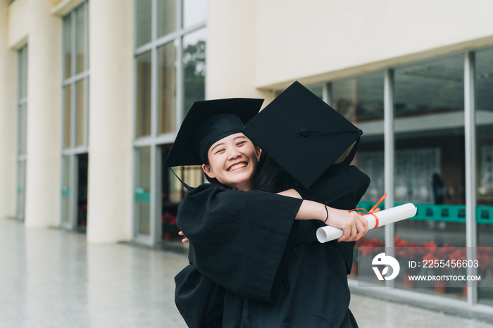 Two asian girls celebrating exam results in school corridor. happy young college student holding diploma hugging best friends in gown and mortarboard on graduation ceremony standing outdoor campus.