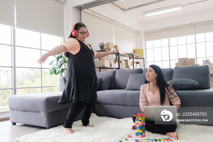 Asian mother using laptop and young girl with autism wearing headphone dancing on the floor at home with his mother. Autistic young students are learning with teachers.