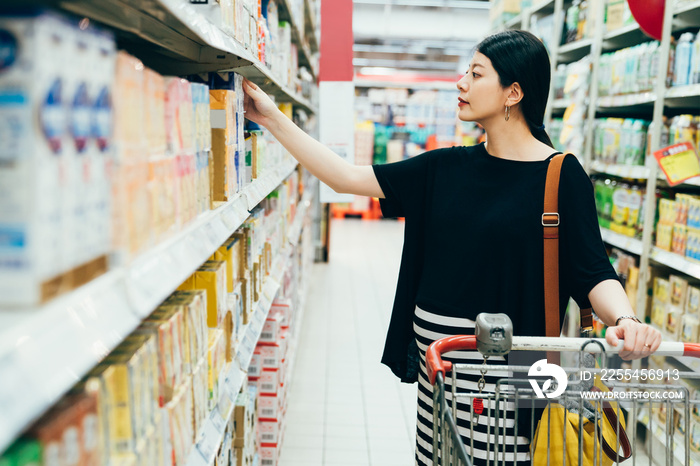 beautiful asian japanese future parent girl take drink from shelves of beverage department of supermarket. pregnant lady shopping in hypermarket. side view motherhood pushing cart in grocery store