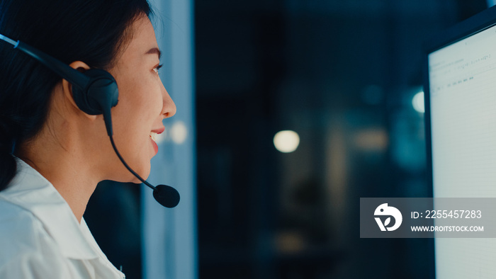 Millennial Asia young call center agent or customer support service executive using computer and microphone headset working technical support in late night office. Telemarketing or sales job concept.