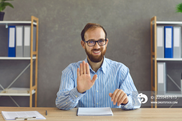 Millennial man with distrustful smile sitting at home office desk looking at camera showing stop palm hand gesture. Displeased young bearded business man refusing offer during online video conference