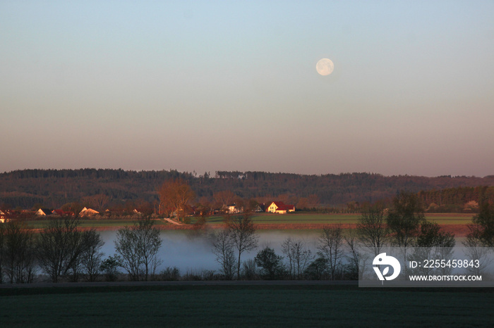 Landscape with fog over Altmühl river valley on a spring morning with pale full moon and Hilsberg village in the background, Franken region in Germany