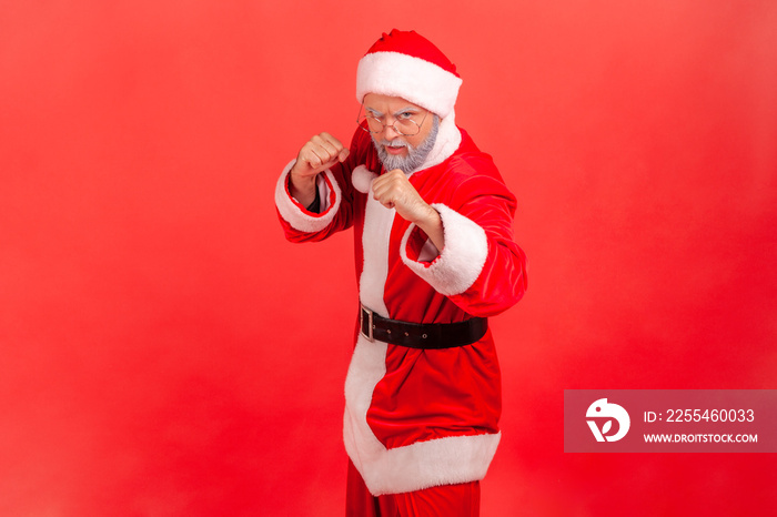 Strict elderly man with gray beard wearing santa claus costume standing with fists and being ready to fight, looking at camera with angry expression. Indoor studio shot isolated on red background.