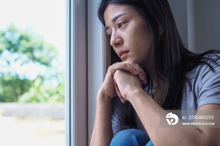 An Asian woman was sadly sitting by the window in the room. Female feels unhappy and disappointment about problem in personal life. Depression or unexpected pregnancy concept