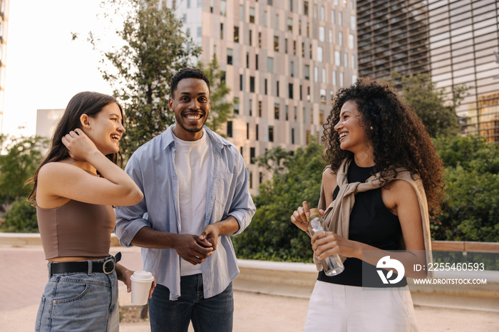 Smiling young caucasian, african friends talking on funny topics standing outdoors. Brunette guy and girls wear casual clothes. Sincere emotions lifestyle concept