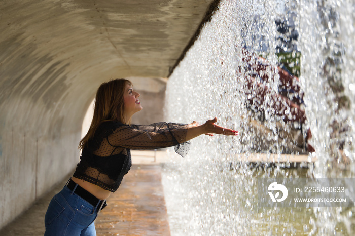 Attractive mature woman in transparent black shirt and jeans, touching the water falling from a waterfall. Concept maturity, beauty, fashion, water, happiness.