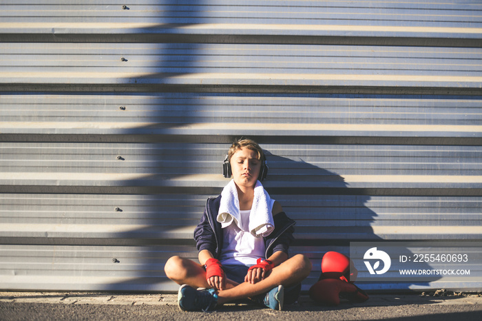 Handsome young male with boxing bands in his hands, relaxing before the match, sitting on the road. Boy ready for a boxe workout listening music. Sport, youth, determination and competition concept.