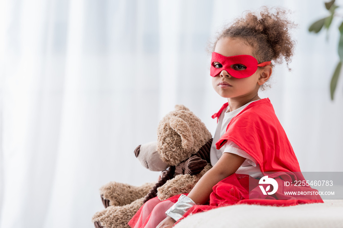 Cute african american child in red superhero costume and mask with teddy bear looking at camera