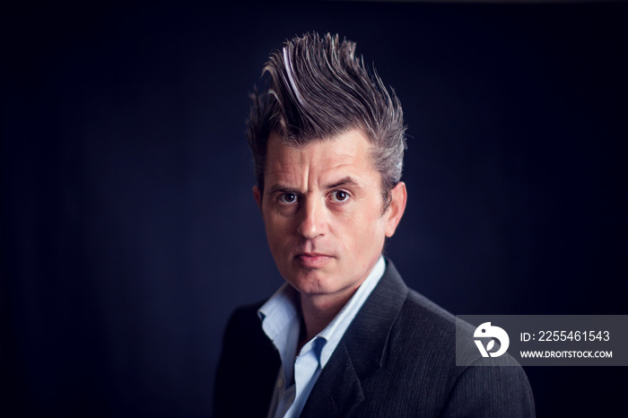 Man with mohawk wearing suit in front of black background.