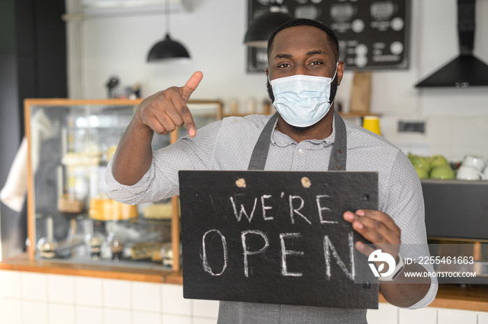 An multiracial waiter, a cafe or bakery owner in protective medical mask is pointing finger at open sign board in his hand. Small business reopening after lockdown