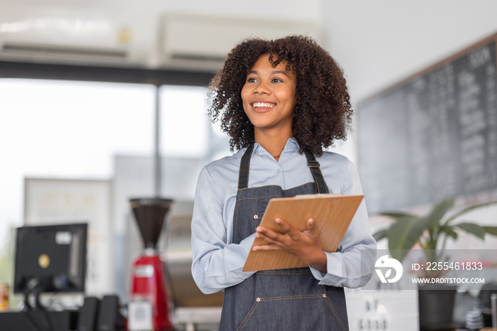 Female African coffee shop small business owner wearing apron standing in front of counter performing stock check. afro hair employee Barista entrepreneur.