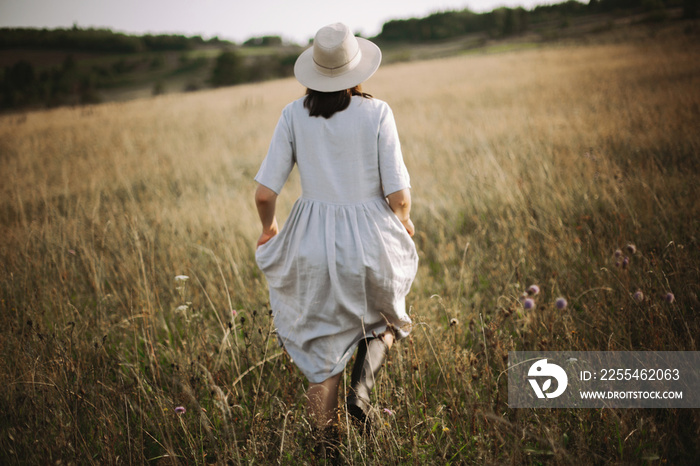 Stylish girl in linen dress running among herbs and wildflowers in sunny meadow in mountains. Boho woman relaxing in countryside, simple rustic life style. Atmospheric image. Space text