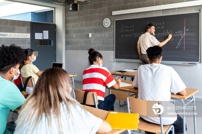 White mature man teacher writes math formulas on blackboard with chalk. Multiracial group of students pay attention to explanation. Copy space.