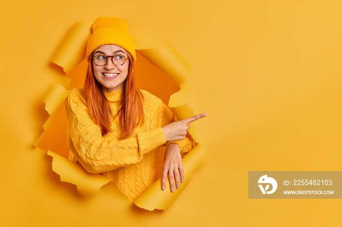 Good looking cheerful millennial girl with tender smiles advertises something suggests discounts indicates at blank space dressed in casual jumper and hat poses in ripped hole of yellow background