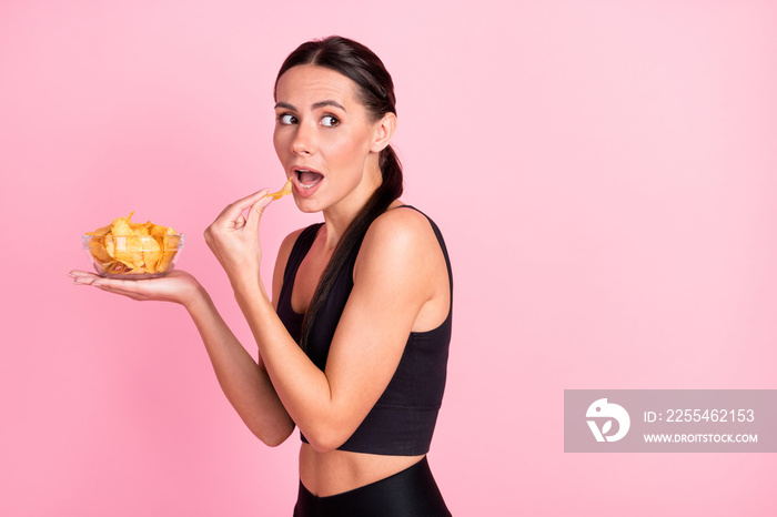 Photo of doubtful hiding young lady sportswear eating potato crisps looking back empty space isolated pink color background