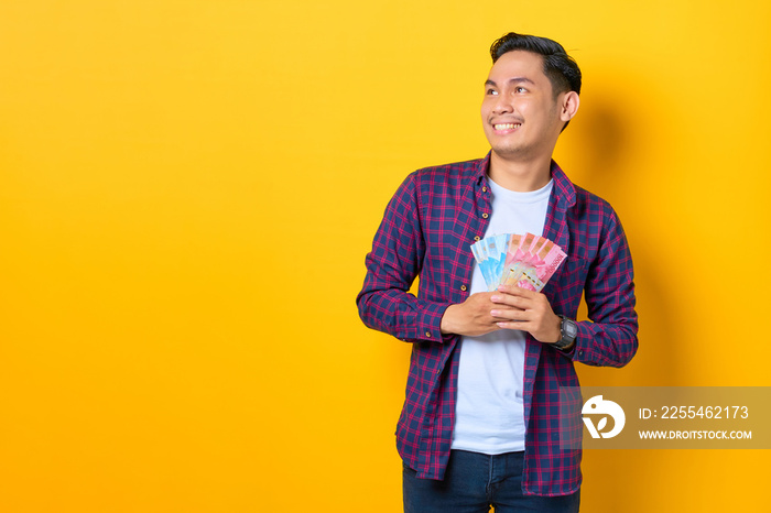 Smiling young Asian man in plaid shirt holding money banknotes and looking away at copy space isolated on yellow background