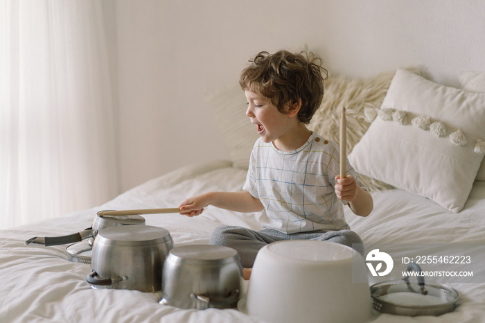 Cute young boy using wooden sticks to bang saucepans that are set up like a drumset.